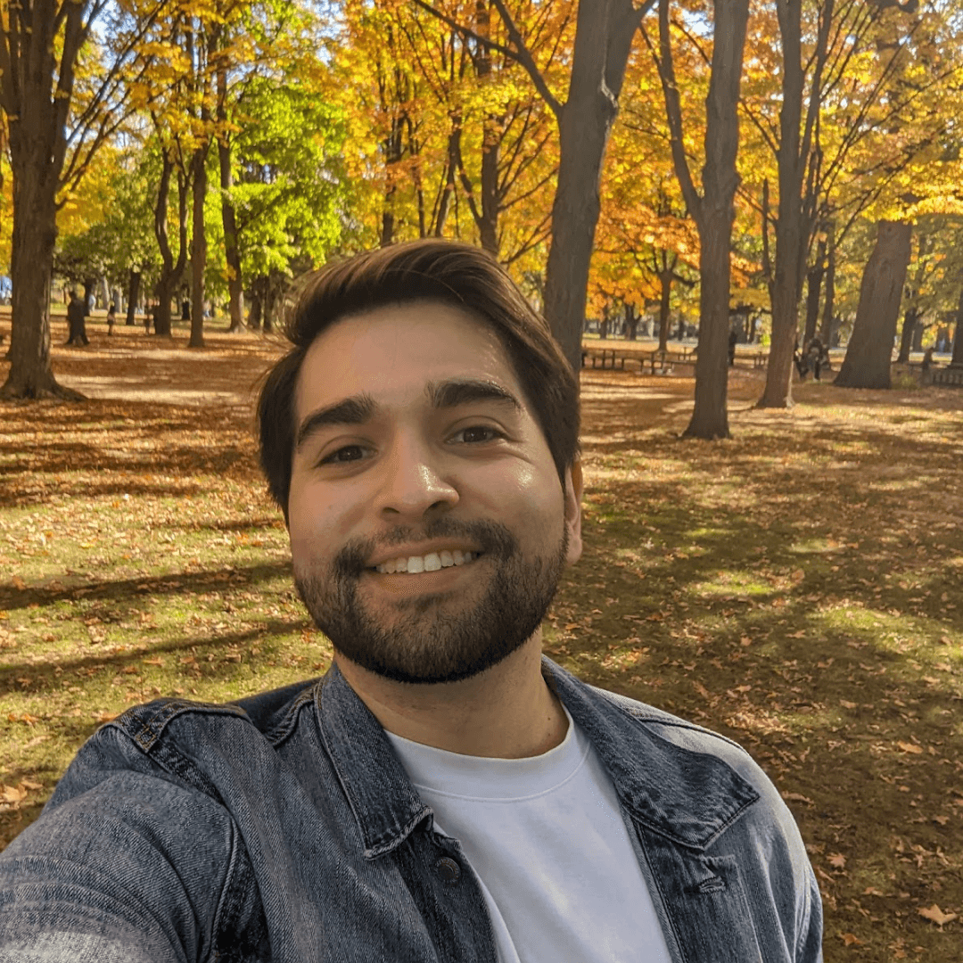 Me - JJ, a smiling person with a beard wearing a jean jacket standing in front of an autumnal orange and yellow forest.